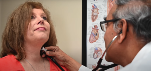 A concerned woman is being examined by a doctor using a stethoscope in a clinical setting, with educational anatomy posters in the background.